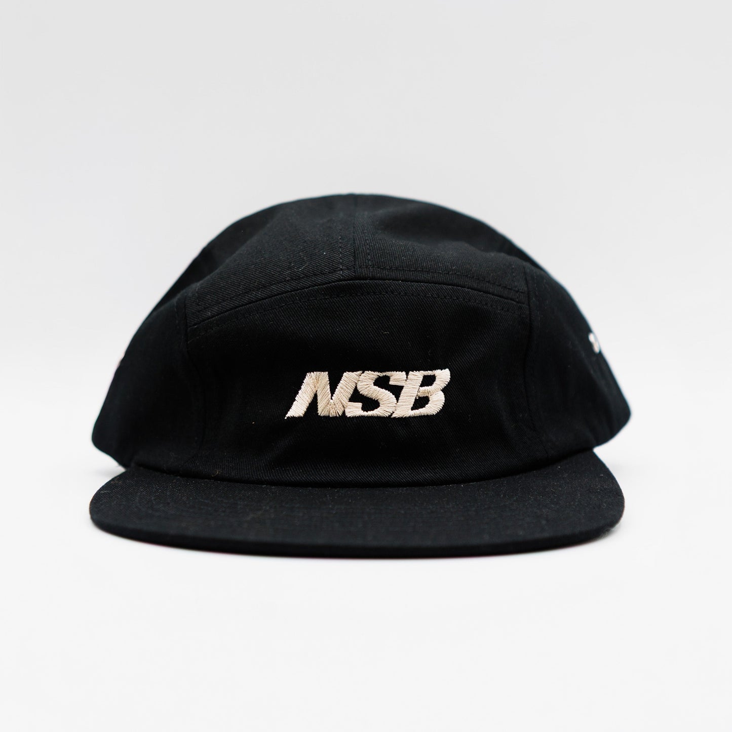 A black five-panel cap with a structured crown and a flat brim, featuring the letters 'MSB' embroidered in metallic white on the front panel, set against a plain light gray background.