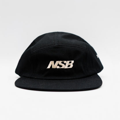 A black five-panel cap with a structured crown and a flat brim, featuring the letters 'MSB' embroidered in metallic white on the front panel, set against a plain light gray background.