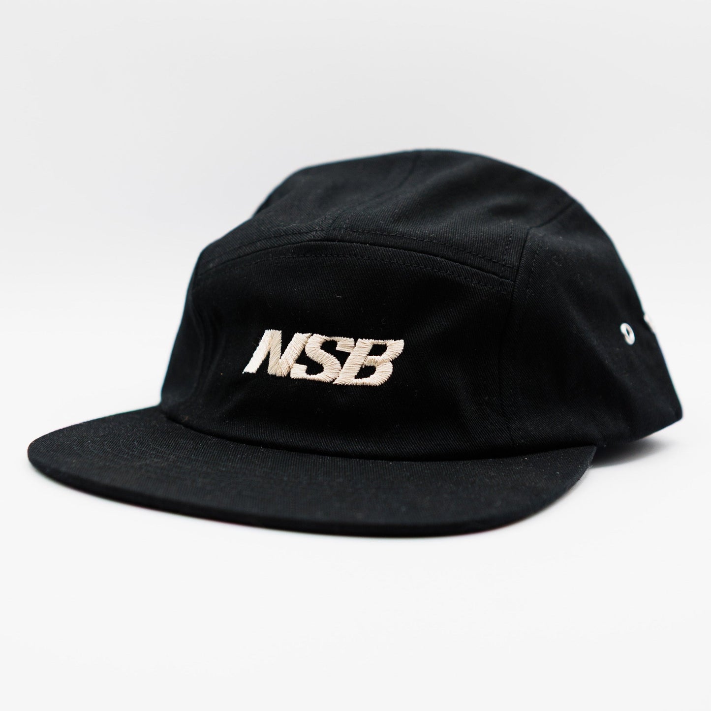 A black five-panel camp cap with a flat brim, featuring the letters 'NSB' embroidered in a metallic white tone on the front, set against a plain white background.