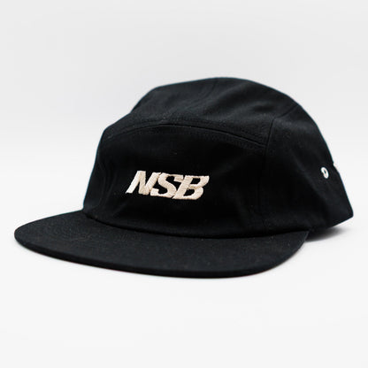 A black five-panel camp cap with a flat brim, featuring the letters 'NSB' embroidered in a metallic white tone on the front, set against a plain white background.