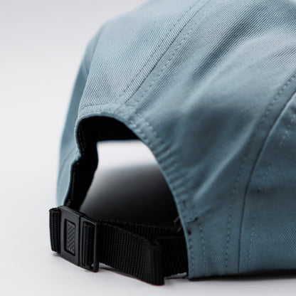 Close-up of the back of a dusty blue fabric cap with a black adjustable strap and plastic buckle, photographed on a light gray studio background.