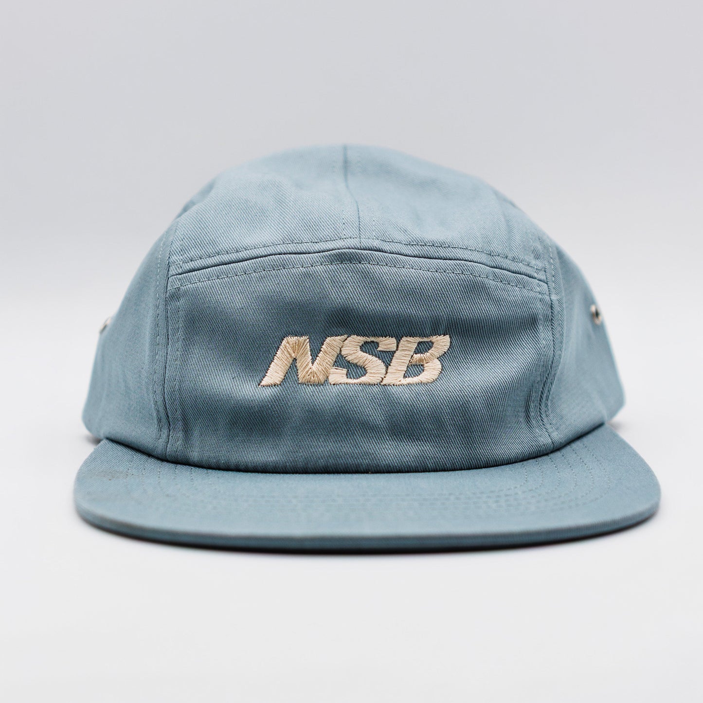 A light blue denim-style 5-panel cap with a flat brim, featuring the brand name 'NSB' embroidered in gold on the front, set against a neutral light gray background.