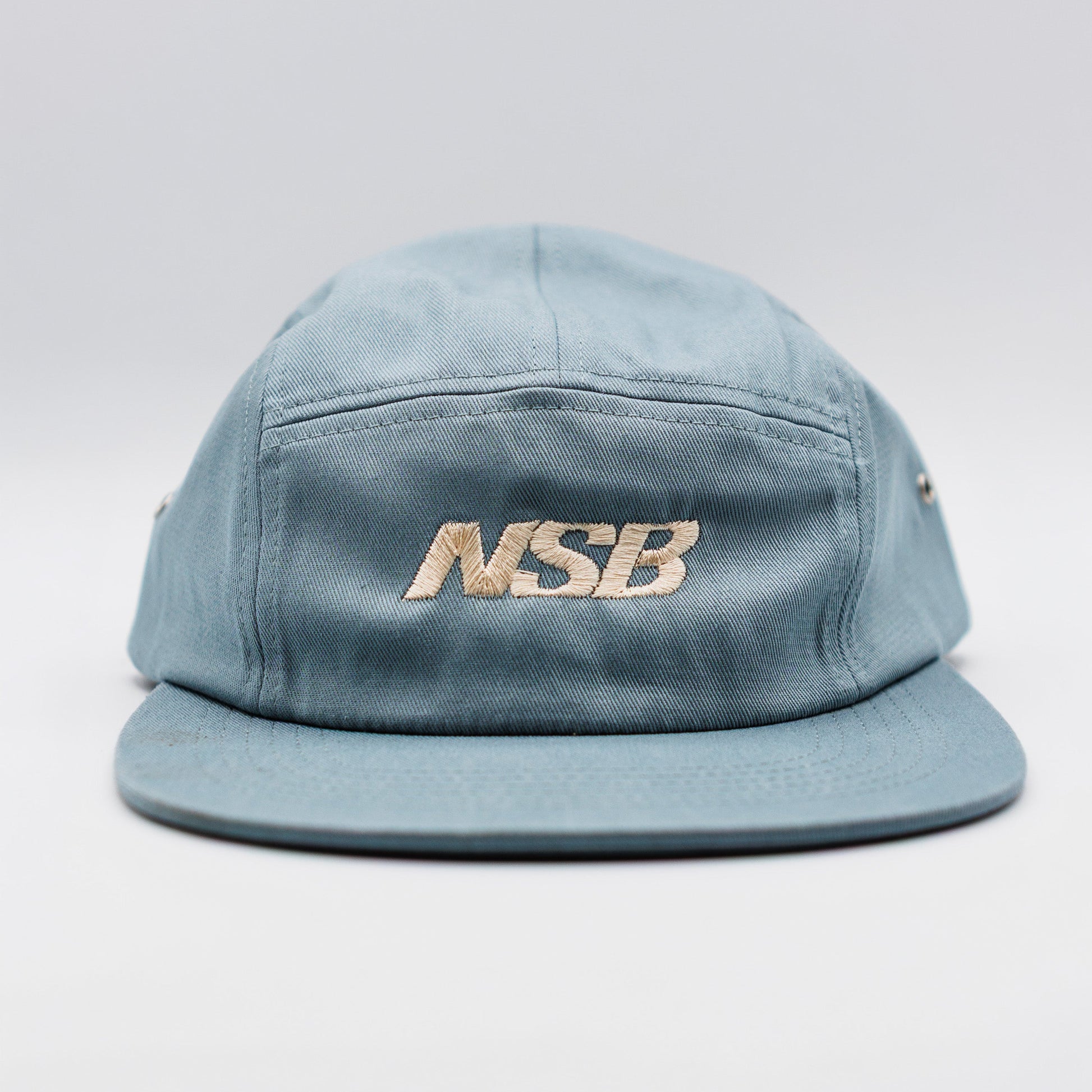 A light blue denim-style 5-panel cap with a flat brim, featuring the brand name 'NSB' embroidered in gold on the front, set against a neutral light gray background.