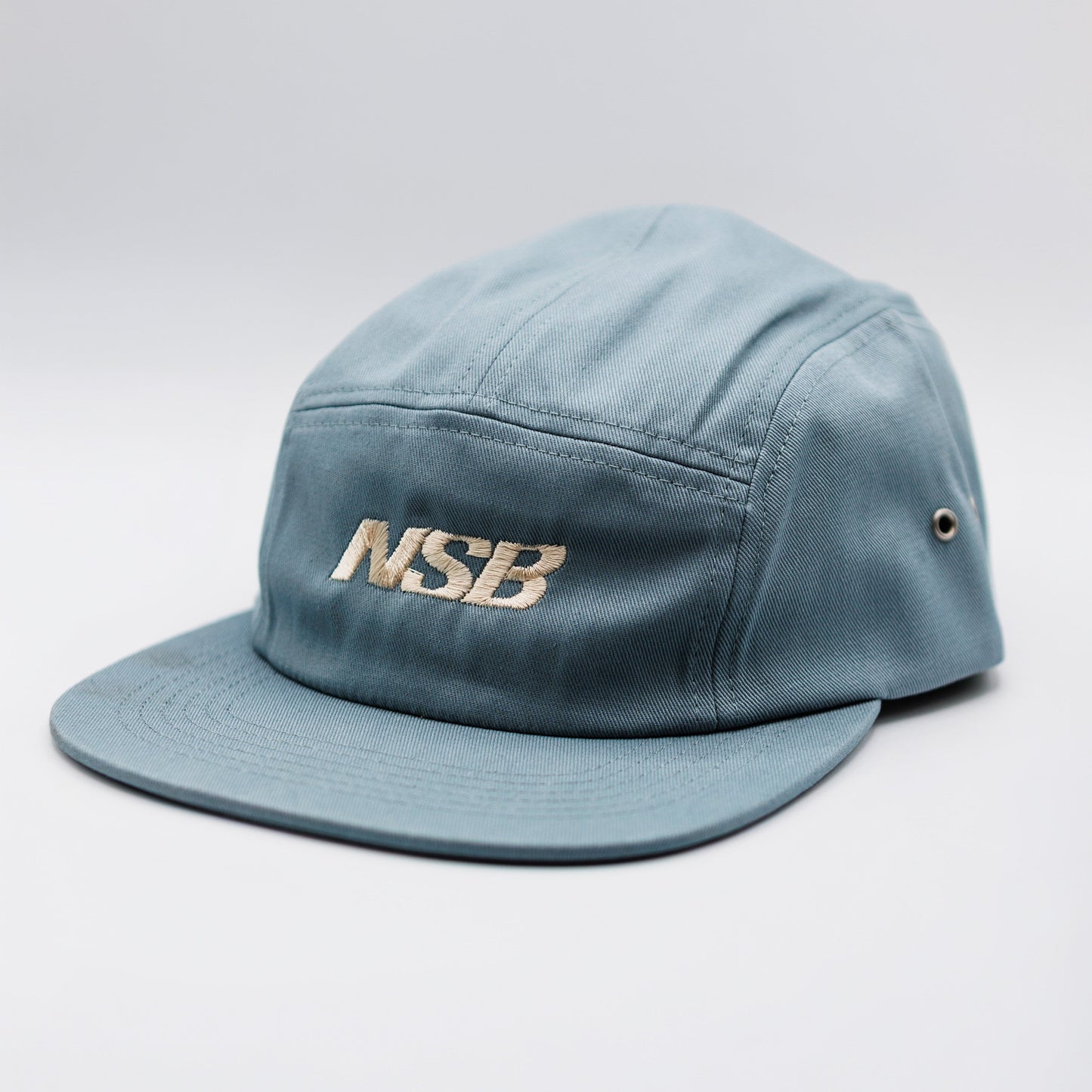 A light blue five-panel camp cap with 'NSB' embroidered in gold on the front, set against a neutral light gray background.