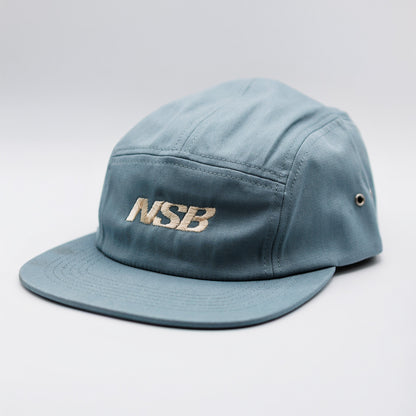 A light blue five-panel camp cap with 'NSB' embroidered in gold on the front, set against a neutral light gray background.